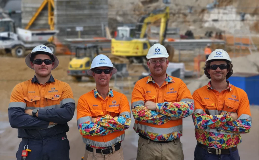 Four construction workers wearing orange safety shirts and helmets stand confidently with arms crossed at a worksite. A bulldozer and construction materials are visible in the background, with the workers smiling and facing the camera.