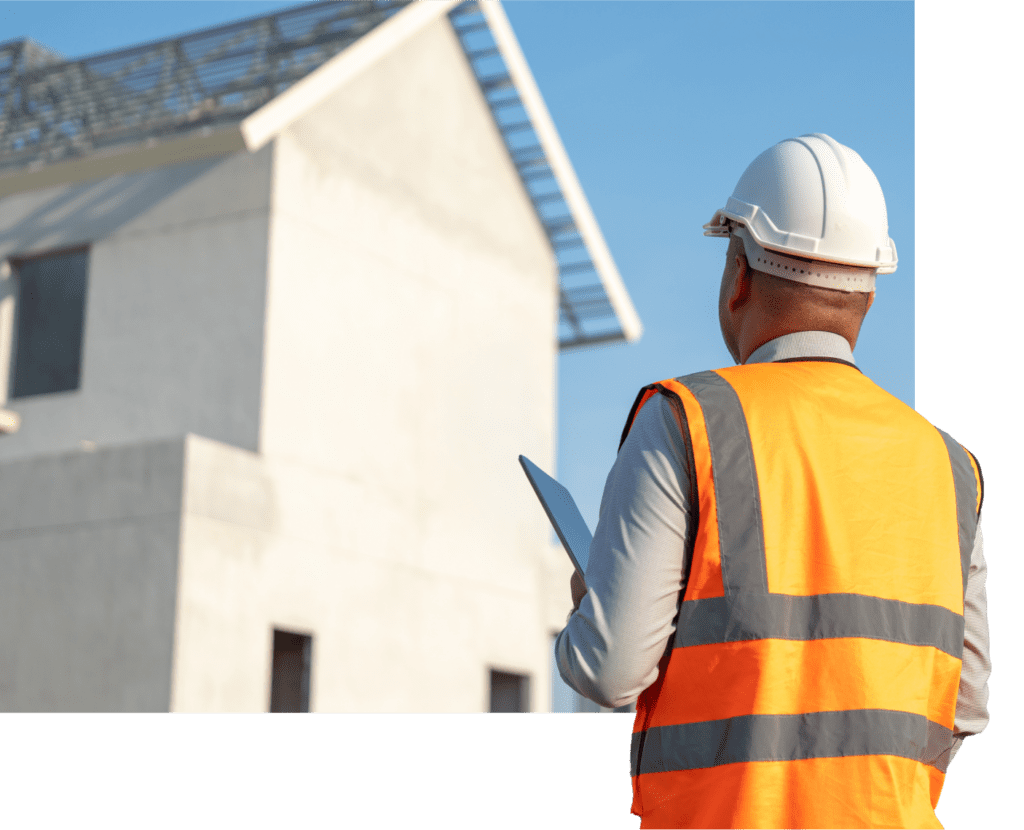 A construction worker in an orange vest and white hard hat holds a tablet, standing in front of a partially constructed building with a slanted roof, against a clear blue sky.