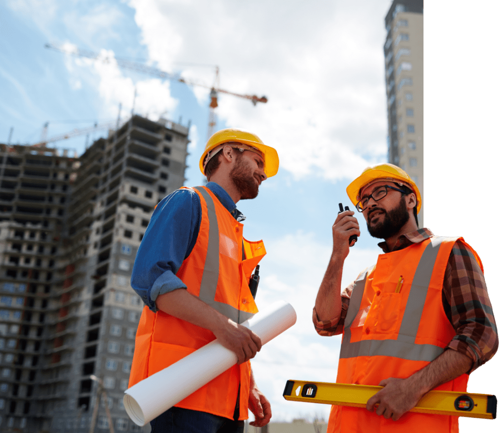 Two construction workers wearing orange safety vests and yellow hard hats stand in front of a building under construction. One holds a rolled-up blueprint, and the other speaks into a walkie-talkie while holding a level tool.