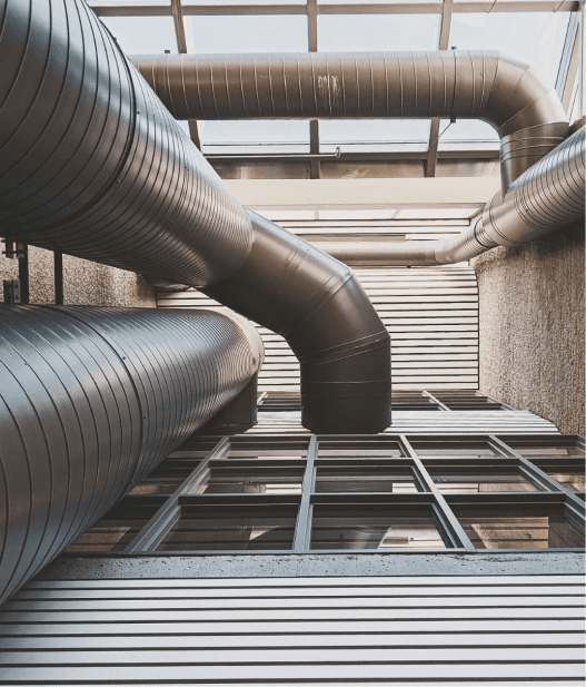 View of large industrial ventilation pipes against a modern building facade with metal and glass elements, creating a geometric pattern along with the ceiling structure. The image captures an urban architectural design.