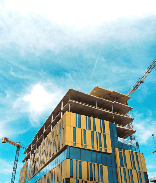 A modern building under construction with a blue and yellow facade, featuring cranes in the background against a bright blue sky with scattered clouds.