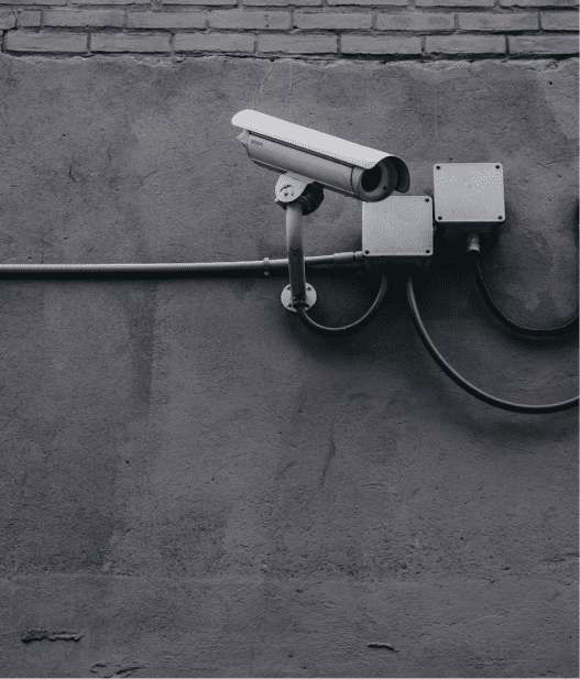 A security camera mounted on a gray concrete wall, surrounded by junction boxes and cables. The camera is facing downward, suggesting surveillance. The wall is slightly textured, with a brick pattern near the top.