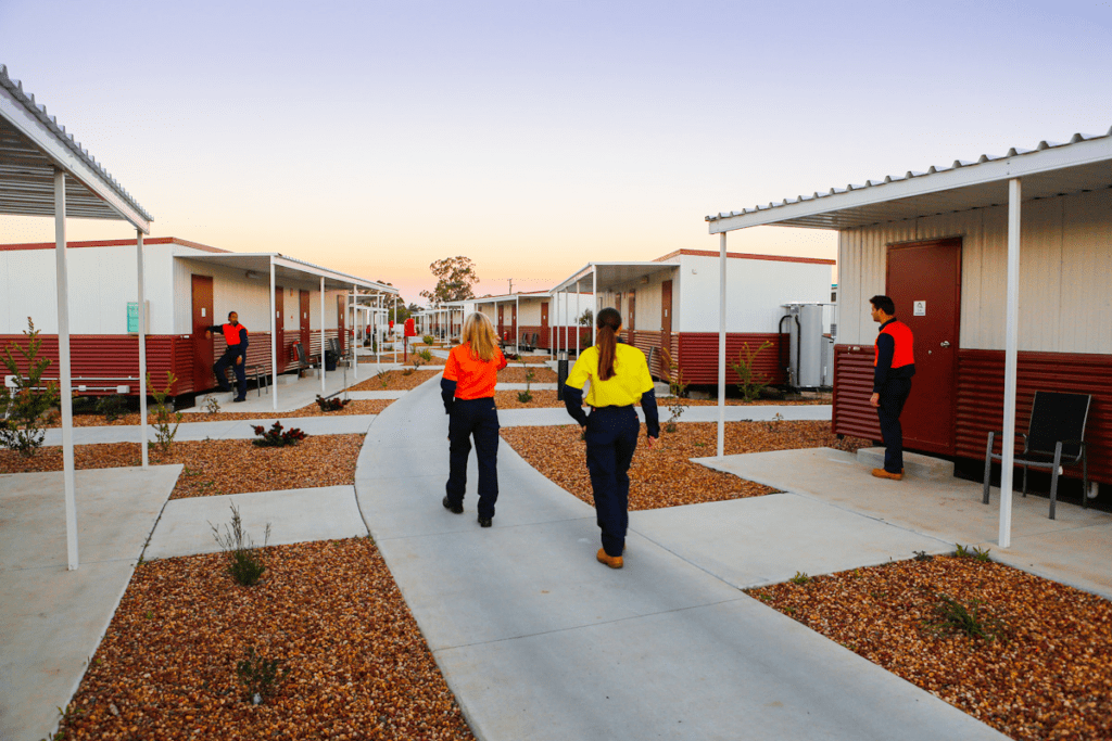 People in colorful uniforms walk between small white and red cabins with landscaped paths at sunset. Some workers are interacting near the buildings, and the sky is clear in the background.