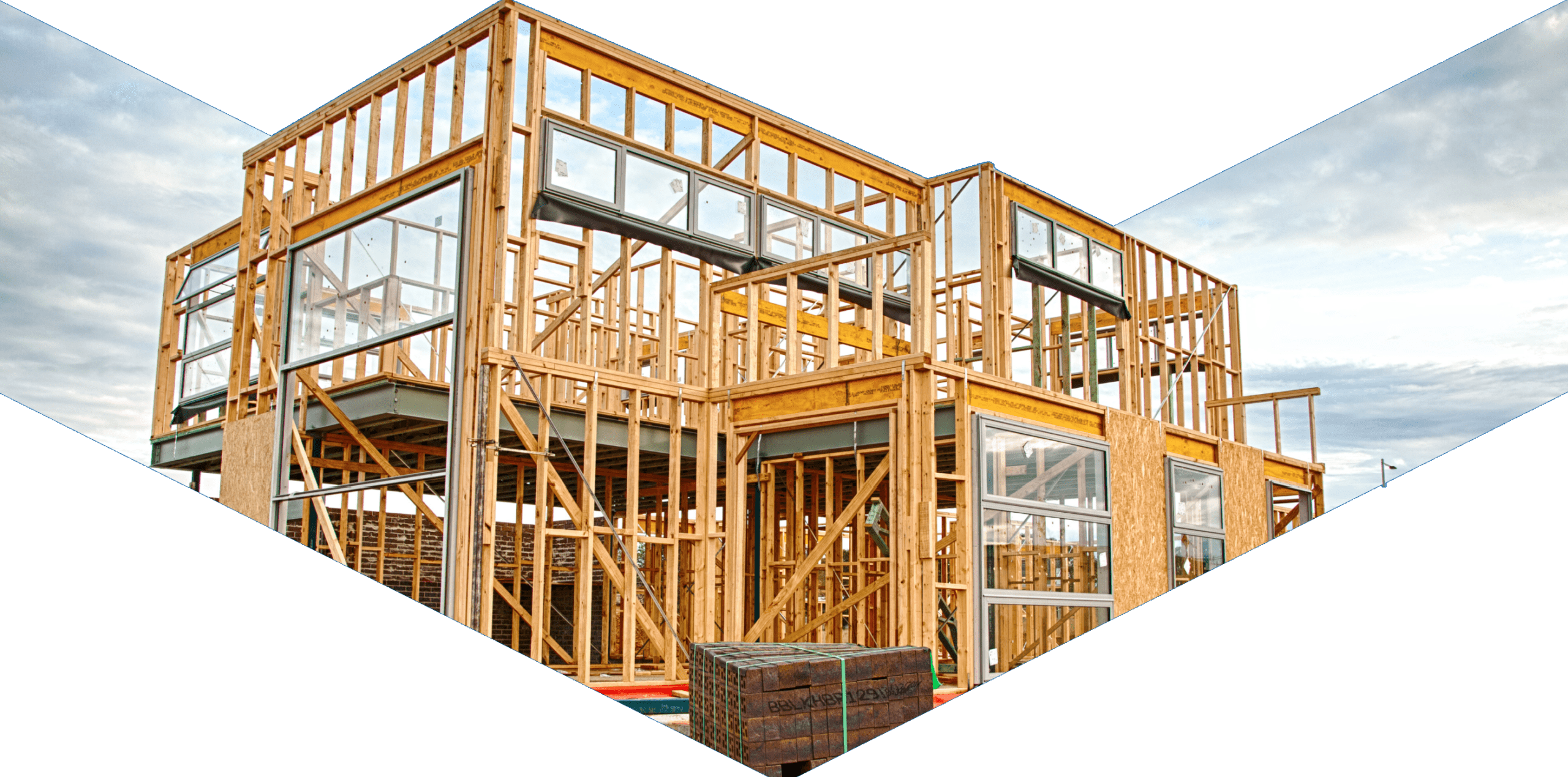 A two-story house under construction, featuring a wooden frame, open walls, and visible windows. The sky is cloudy in the background, and building materials are stacked nearby.